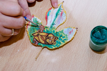 A grown woman is painting with paint on an autumnal tree leaf. Hands paint a funny bear cub sitting on a log in the woods. Part of a series. Creativity, art.の写真素材