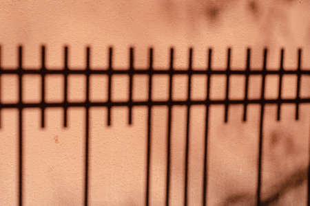 Shadows of a fence and tree branches on an uneven coral back wall. Dark shadows of the metal fence bars. Decorative plastering of the building exterior. Selective focus. No people.の写真素材