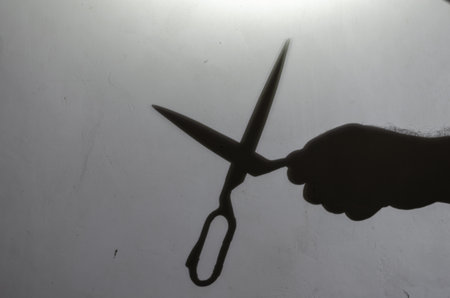 Blur, defocus, noise, grain effect. Dark silhouette of a man's hand with open scissors on a light gray wall. Man holding large scissors. Inside the room. Selective focus.の写真素材