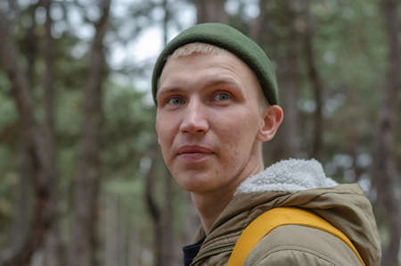 Portrait of a young adult male walking in the woods. Male wearing a green jacket and hat with a yellow backpack behind him. Selective focus.の写真素材