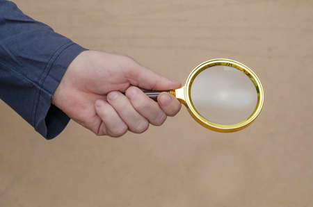 An adult man holds a magnifying glass in his hand against a brown background. A large round magnifying glass in a gold frame. Close-up. Selective focus.の写真素材