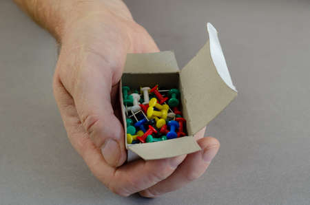 Right Hand and push pins on gray background. A man is holding a box of multicolored thumbtacks. Close-up. Selective focus.の写真素材