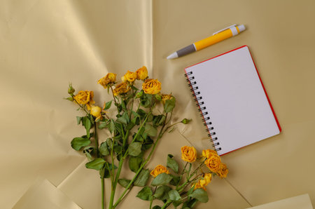 Faded flowers and a blank open notebook with a pen against a creamy surface. Yellow roses. Rectangular spring-loaded notepad. Crumpled shiny wrapping paper. Top view. Selective focus.の写真素材
