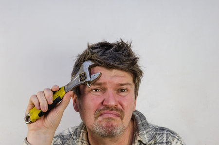 An adult man with disheveled, tangled hair puts a wrench to his forehead. The man is experiencing a headache. Indoors. Selective Focus.の写真素材