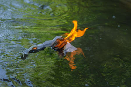 A burning piece of white paper floats down the river. A crumpled business document is engulfed in flames. blurred motion. selective focus.の写真素材