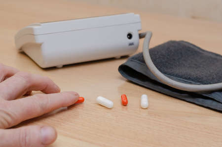 A man's hand takes colorful pills from a table. Digital blood pressure monitor in the background. Checking blood pressure at home. Cardiology. selective focus.の写真素材