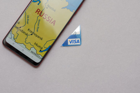 Smartphone and Visa Bank Card Piece against a gray background. There is a map of Russia on the smartphone display. A cut-off piece of blue credit card. Selective focus. Kiev, Ukraine - 01 05 2022.のeditorial素材