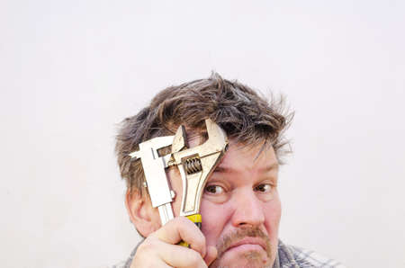 A tired Adult male puts a metal hand tool to his forehead. Holds a caliper and a wrench. A man with tangled, disheveled brown hair against a light gray wall. indoors. selective focus.の写真素材