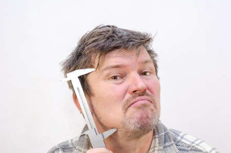 The male grimaces, measuring his face with a caliper. A man with tangled, disheveled hair in front of a light gray wall. indoors. selective focus.の写真素材