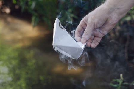 A man's hand holds a smoking paper boat. A white ship and puffs of smoke against a dark green background. Blurred, defocus, selective focus, noise, grain effectの写真素材