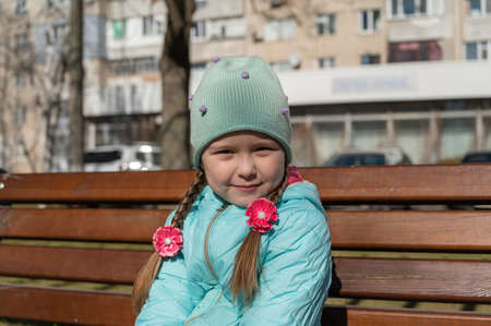 Portrait of a 5-year-old girl sitting on a park bench. Shy girl with long hair braided into pigtails. A child in a blue jacket and hat. springtime.の写真素材