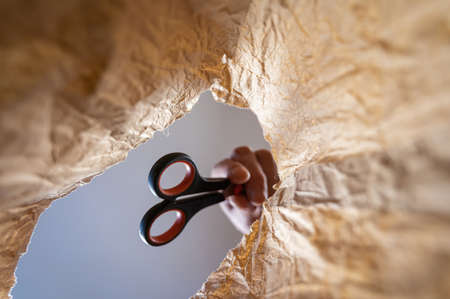 A hand places scissors in a paper bag. A mature man's hand holds office scissors over an open brown bag. Shot from the bottom up. close up. selective focus.の写真素材