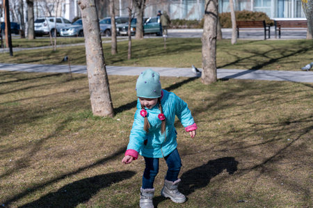 A girl is walking in a spring city park. A five-year-old child in a blue jacket with pink cuffs...