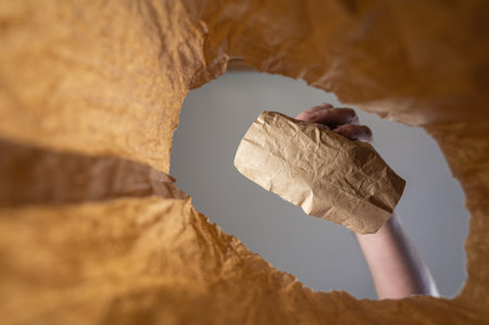 A hand places a paper-wrapped lunch in a paper bag. A mature man's hand holds the food with him over...