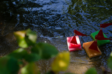 The view through the green shoreline foliage of children's ships floating on the river. Colorful...