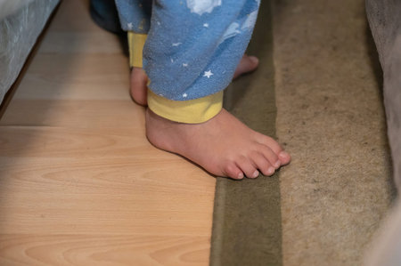 Bottom section of a child standing barefoot on the bedroom floor. A five-year-old boy in blue pajamas is standing next to his bed on the carpeted floor.の写真素材