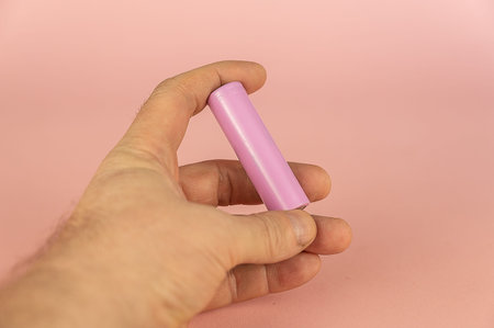 An adult male hand holding a lithium-ion battery against a pink background. The cylindrical...