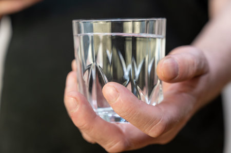 A close-up of a hand holding a glass of water. A mature man in a black shirt holds a glass of...