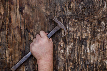 A man's left hand holds an old iron hammer against a dark wooden background. Cracked wooden surface with cracks and chips. selective focus.の写真素材