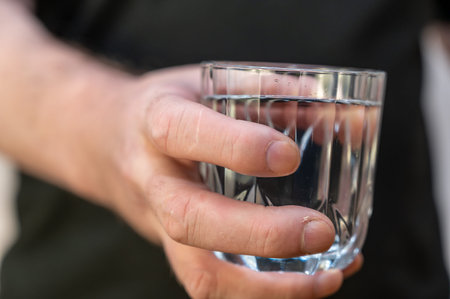 A close-up of a hand holding a glass of water. A mature man in a black shirt holds a glass of...