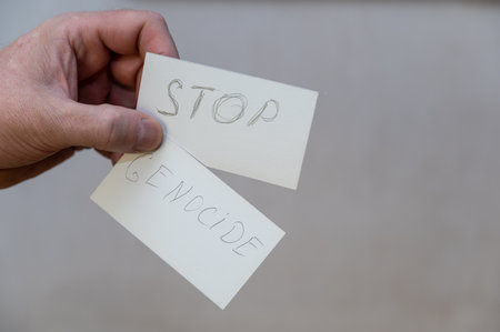 Stop Genocide. Words written in jagged letters. A man's hand holds two white paper rectangles with text against a gray background.の写真素材
