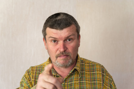Portrait of an adult man in a plaid shirt against a beige background. Man with a short haircut and graying hair points his index finger at the camera. front view. indoors.の写真素材