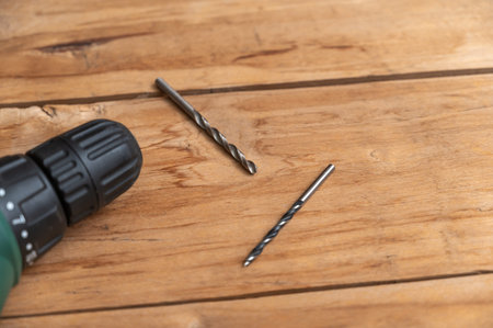 An electric drill and two drills lie against the wooden surface. The hand-held power tool is turning...
