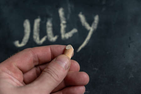 Adult male holding a piece of yellow chalk in his hand. Handwritten word JULY on black chalkboard. selective focus.の写真素材