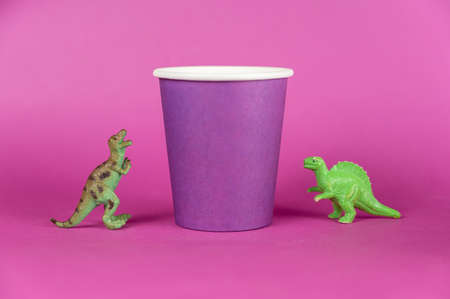 Dinosaur miniatures and a paper cup against a purple background. A lilac disposable drink glass. Green figures of extinct animals of prey standing on their hind legs. selective focus.の写真素材