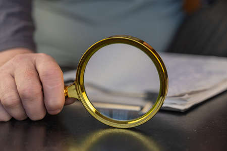 Close-up of a man's hand holding a magnifying glass. Round magnifying glass with gold-colored frame. paper clipboard background. selective focus.の写真素材
