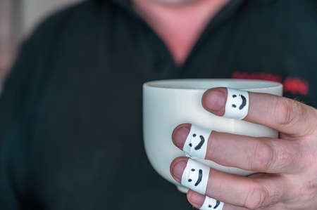 Ceramic cup in a man's hands. The first phalanges of the fingers are wrapped in a white ribbon. Smiling faces are painted on the bandages. positive emotion concept. selective focus. close-upの写真素材