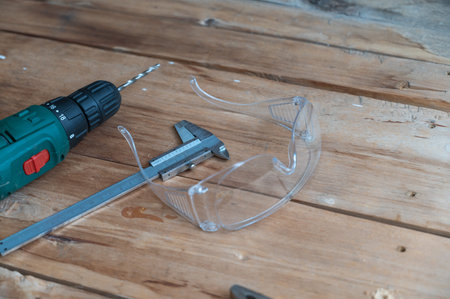 Electric drill, caliper, pliers and safety goggles lie on a wooden surface. The drill is mounted in...