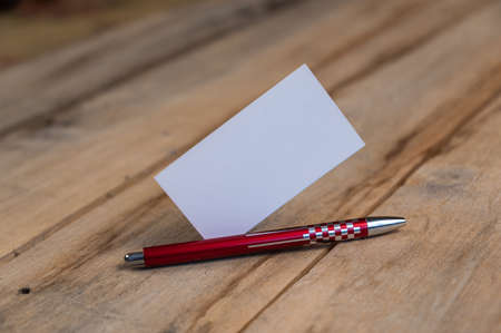 A blank business card and a red pen against a wooden background. A white paper rectangle and a ballpoint pen lie on top of old cracked boards. selective focus.の写真素材