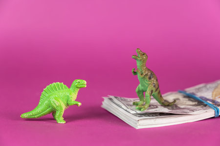Dinosaur miniatures and a wad of hundred-dollar bills against a purple background. A stack of money...