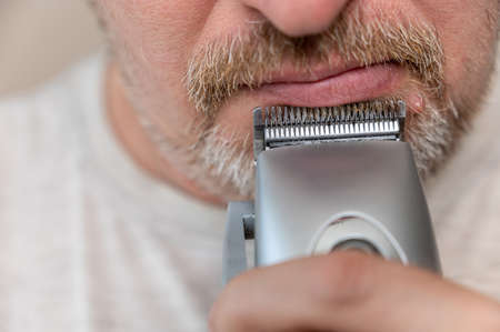A mature man shaving. The bottom of a man's face. Man cuts facial hair with a hair clipper. A male with stubble. Gray hair on his beard and mustache. close-up. selective focus.の写真素材
