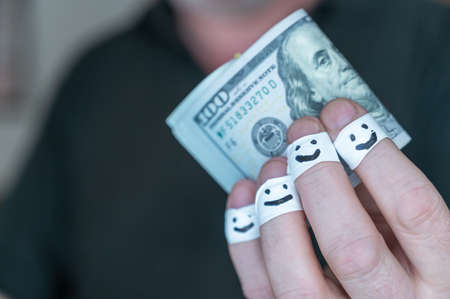 An adult man holds out his hand with $100 bills to the camera. The first phalanges of his fingers are wrapped in white ribbon. Smiling faces are painted on the bandages. positive emotion concept.の写真素材