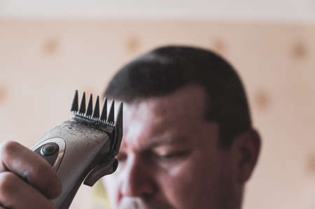 A middle-aged man cuts his own hair. The person is holding an electric haircutting machine in front of his head. Inside a living room. front focusの写真素材