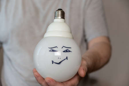 A middle-aged man is holding an electric lamp with his face painted on it. A happy smiling face is drawn in black marker. A large round electric lamp in the man's hand. Middle section of a mature manの写真素材