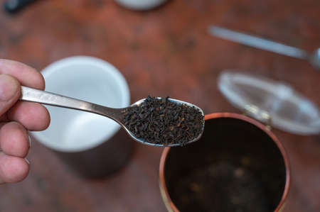 Middle-aged man holding a spoonful of dry tea. Crushed black tea leaves in spoon over red table. Cup and container are blurred on background. Healthy lifestyle. first person view. front focus. indoor.の写真素材