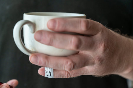 A man is holding a ceramic cup in his hand. The first phalanx of his little finger is wrapped in...