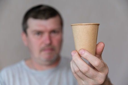 A middle-aged man holds a brown glass in front of his face. A disposable paper drink glass in his...
