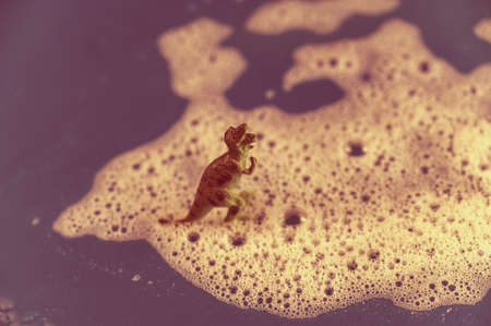 Dinosaur figurine stands in soapy foam. Predator standing on its hind legs with its mouth open. Bubbles of white detergent foam on the floral surface. Shampoo foam texture. selective focus. Tonedの写真素材