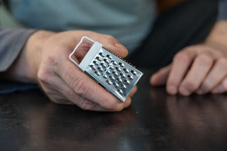 A grown man is holding a small kitchen grater in his hands. A cone-shaped metal grater with a white handle. Man sitting at a black table indoors. close-up. selective focus.の写真素材