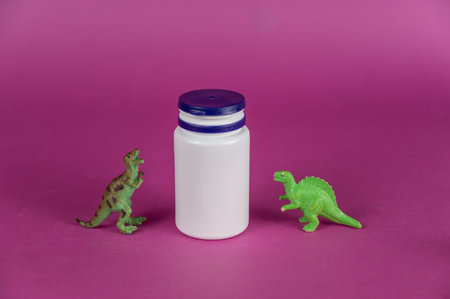 White vial and dinosaur miniatures against a purple background. Small green figures of predatory animals standing on their hind legs. Plastic cylindrical container with blue lid.の写真素材