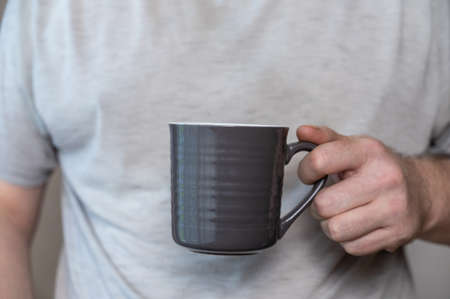 A middle-aged man is holding a gray mug. A ceramic cup with a handle for hot drinks in a man's hand. Middle section of a mature man in a gray T-shirt. inside the room. selective focus.の写真素材