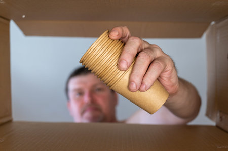 A man pulls glasses out of a cardboard moving box. A set of disposable paper beverage glasses....
