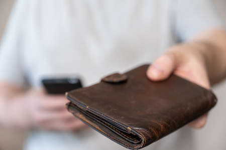 A middle-aged man is holding an old purse. Brown leather wallet in a man's hand. Middle section of a mature man in a gray T-shirt. inside the room. selective focus.の写真素材