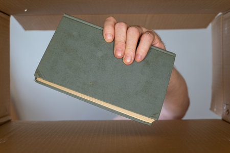 A man's hand pulls a book out of a cardboard moving box. An old green hardcover book. bottom view. inside view. close-up. selective focus.の写真素材