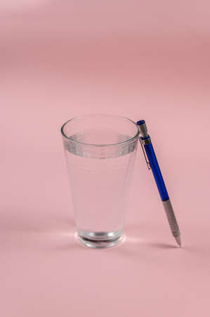 A glass of water and a pencil on a pink background. A full glass of drinking water. Blue and silver metallic pencil. Kurp plan. selective focus.の写真素材