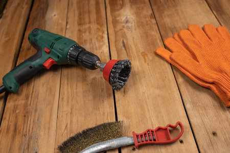 Electric drill, knot cup wire brush, metal brush and orange gloves. Hand and power tools lie on the...